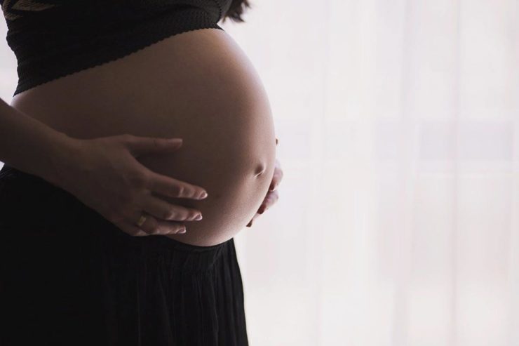 A pregnant woman is holding her belly in front of the window.