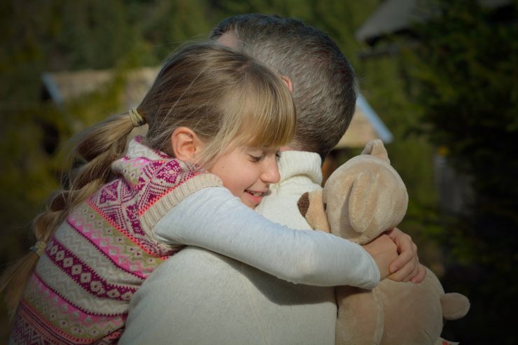 A little girl hugging her father and holding onto his teddy bear.